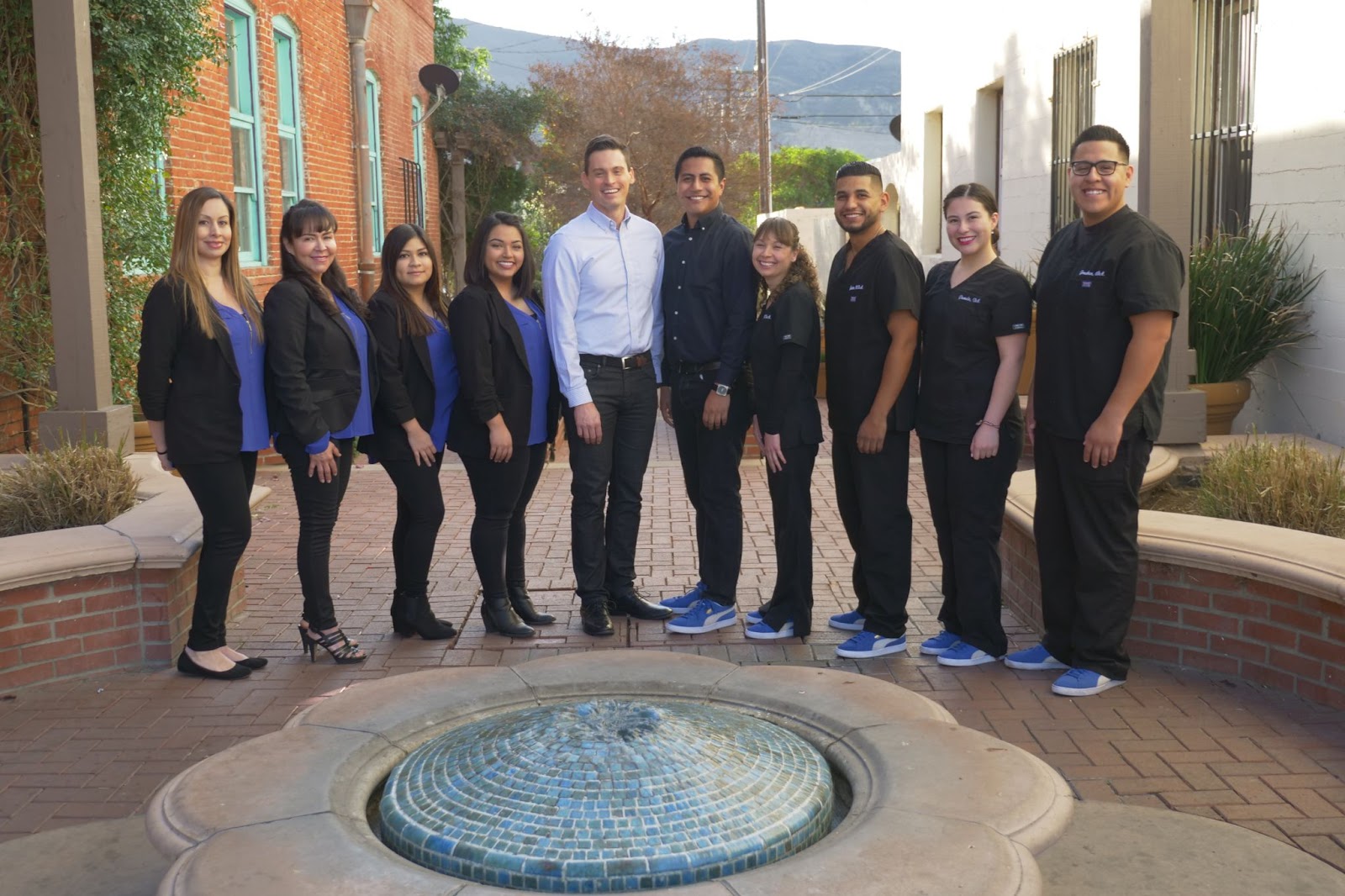 Team of orthodontic professionals from Ventura County Orthodontics, smiling in a casual outdoor setting near a fountain, wearing black uniforms with blue accents, promoting braces-friendly treats in Oxnard.
